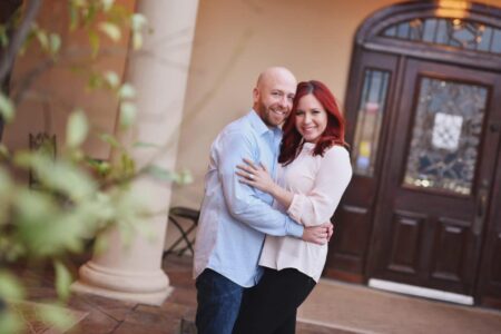 Smiling couple embraces outside a building with a dark wooden door; man in blue shirt, woman with red hair in blouse and black pants.
