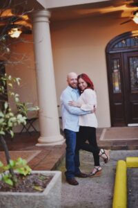 Smiling couple stands by a columned building; woman with red hair in light top, bald man in light blue shirt.