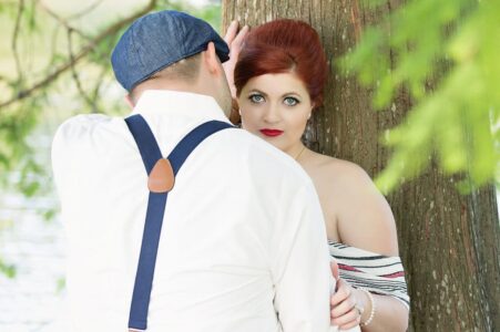 Red-haired woman leans on a tree, looking at the camera; man in white shirt and cap stands with his back to the lens.