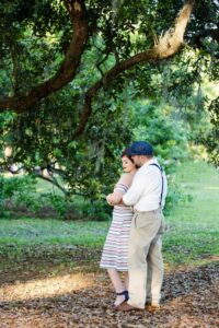 Couple embracing under a large tree in sunlight; woman in striped dress, man in cap, white shirt, suspenders, and khaki pants.