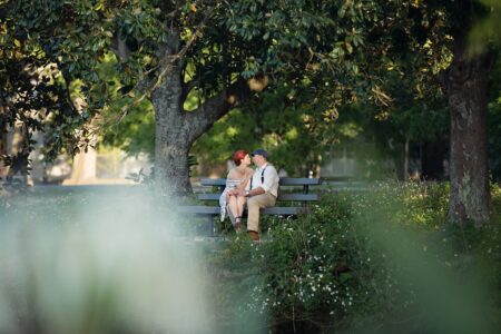 Couple sitting close on a park bench under large trees, surrounded by greenery and soft light, sharing an intimate moment.