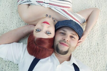 Red-haired woman and bearded man lying on a cream blanket, heads touching, looking up. She wears bold makeup; he has a cap.