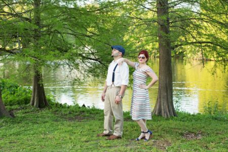 Man in suspenders and cap stands by lake with woman in striped dress; both look away, tall trees behind them.