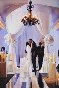 Bride and groom kiss at the altar with white drapes, candles, flowers, and a black chandelier in an elegant indoor wedding.