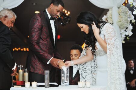 A couple and child pour sand into a vase at a wedding, joined by an older man, with white flowers and drapery as decor.