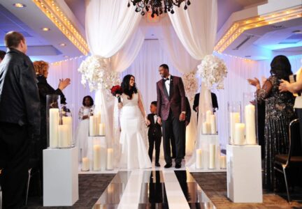 Bride and groom walk with a young boy down the aisle at an indoor wedding, surrounded by guests, candles, and white flowers.