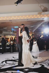 Bride and groom’s first dance on black-and-white floor, watched by guests in black and a little girl, with soft lighting.