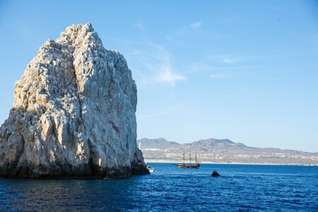 Rocky formation in blue sea with a sailboat and coastal mountains under a clear sky in the background.