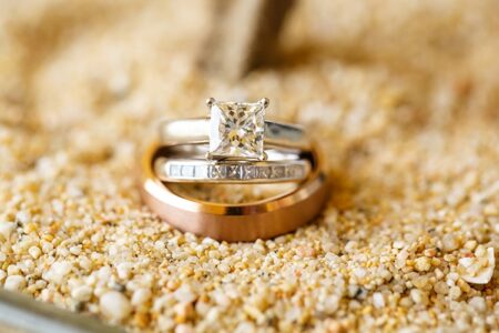 Diamond engagement ring and gold and silver wedding bands on light-colored coarse sand.