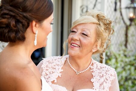 Bride and older woman in elegant attire smile at each other outdoors; the woman wears a pink lace outfit and pearl necklace.