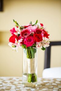 Glass vase with red roses, pink gerbera daisies, and white lilies with greenery on a mosaic-patterned table.