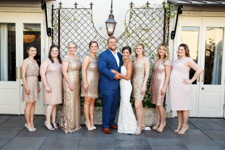 Bride and groom with eight bridesmaids in champagne and gold dresses pose outside a white building with ironwork and plants.
