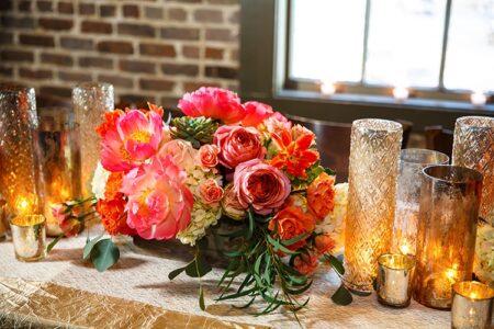 Colorful floral centerpiece with pink, orange, and white blooms, candles, and glass holders on a table by a brick wall and window.