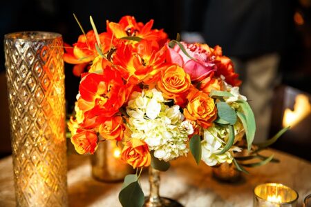 Colorful orange, pink, and white floral arrangement with green leaves sits by a tall gold candle holder on a warmly lit table.