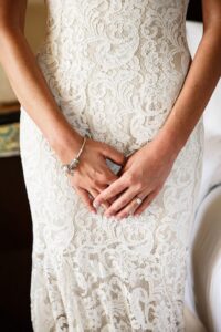Woman in white lace wedding dress with hands folded, displaying a silver bracelet and a ring.