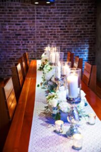 Wooden table with patterned runner, white flowers, and tall glass candles set against a brick wall.