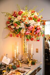 Glass vase with pink, white, and orange flowers, gold candle holders, and lit candles on a white table.
