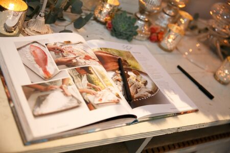 Open cookbook with fish recipe and step-by-step photos, pen on page, cozy decor with candles and flowers.