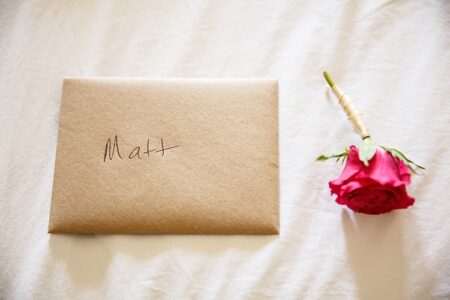 Brown envelope labeled “Matt” on a white surface beside a red rose with its stem wrapped.