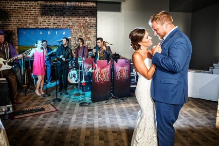 Bride and groom share their first dance as a live band plays; guests watch on a warmly lit dance floor.