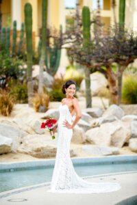 Bride in white lace gown with red bouquet smiles by pool; desert plants, rocks, and tall cacti in the background.