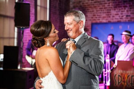 Bride in strapless dress dances with smiling older man in gray suit; live band plays in background.