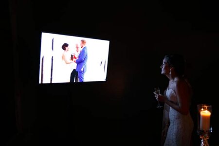 Bride in white dress holds champagne and watches wedding ceremony on screen in dim, candlelit room.