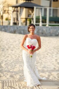Bride in white lace wedding dress smiles on sandy beach, holding pink bouquet; gazebo and building blurred behind.