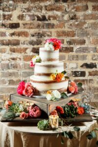 Three-tier naked cake with colorful flowers on a rustic stand, surrounded by candles and succulents against a brick wall.