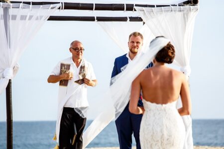 Bride in white dress walks to groom and officiant under white arch on beach, ocean in background.