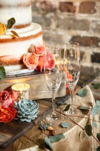 Champagne flutes and a frosted cake with roses, succulent, candles, and eucalyptus on a gold tablecloth before a brick wall.