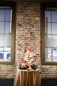 Tiered white wedding cake with colorful flowers on a gold table, framed by brick walls, windows, candles, and a street view.
