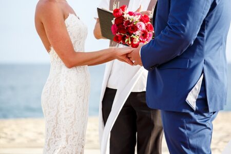 Bride in lace dress and groom in blue suit hold hands at beach wedding with officiant and red-pink flower bouquet.