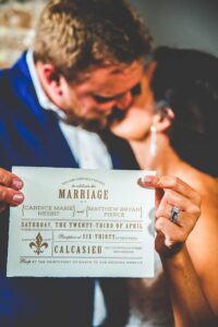 Bride and groom kiss, holding a clear wedding invitation with visible ring; couple softly blurred in background.