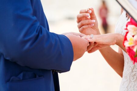 Couple exchanging wedding rings, one in a blue suit and the other in a white dress holding flowers.