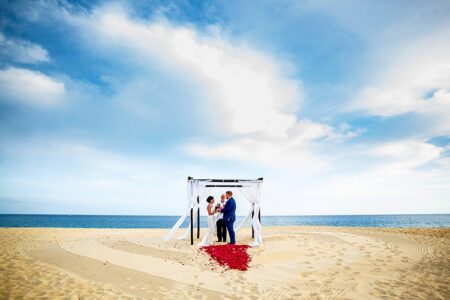 Couple exchanging vows under a white canopy on a sandy beach with rose petals, ocean, and blue sky in the background.