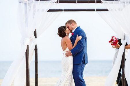 Bride and groom kiss under a white beach canopy with ocean behind them; a person holding flowers stands nearby.