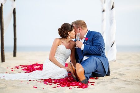 Bride and groom kiss on rose petals under a white canopy at the beach, holding a glass with the ocean behind them.