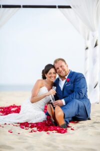 Bride in white dress and groom in blue suit sit on sandy beach holding hands under a canopy, rose petals around them.