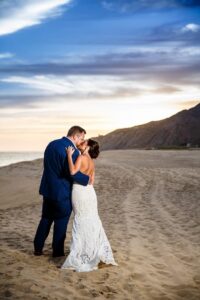 Bride and groom kiss on a sandy beach at sunset, mountains behind them under a vibrant, colorful sky.