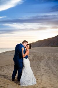 Bride and groom smiling and embracing on a sandy beach at sunset, ocean and cliffs in the background.