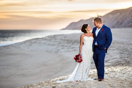 Bride in white lace gown with red flowers and groom in blue suit smile on sandy beach at sunset, cliffs and ocean behind.