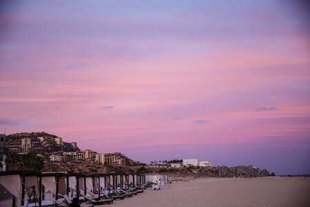 Sunset on a sandy beach with empty lounge chairs, cabanas, resorts, and rocky cliffs under a pastel sky.