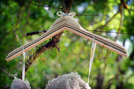 Elegant pearl hanger with “Mrs. Laurant” in gold wire hangs on a tree, lacy garment behind, under green leaves.