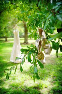 White high heels hang from a tree branch outdoors, wedding dress blurred in the background at a New Orleans ceremony.