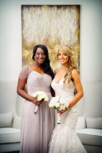 Two women in formal dresses hold bouquets and smile at a New Orleans wedding, posing indoors before a gold abstract painting.