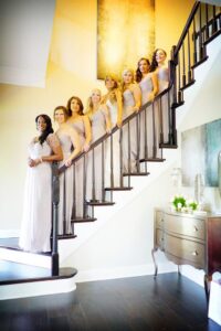 Seven women in matching gray dresses smile on a wooden staircase in a bright, elegant, modern home—New Orleans wedding inspiration.