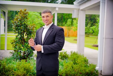 Man in black suit and bow tie adjusts cufflinks outdoors, smiling amid greenery and modern architecture—stylish wedding inspiration.