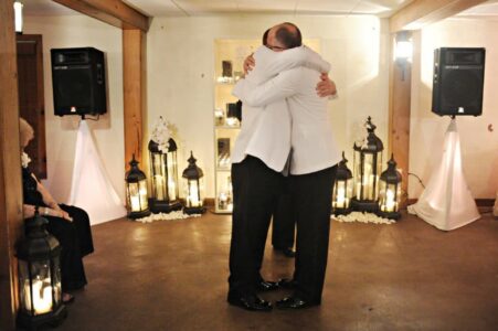 Two men in white jackets embrace in a warmly lit, wedding-inspired room as an older woman looks on.