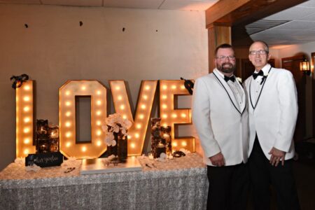 Two men in white tuxedos smile together before a large, lit LOVE sign at an indoor New Orleans wedding.
